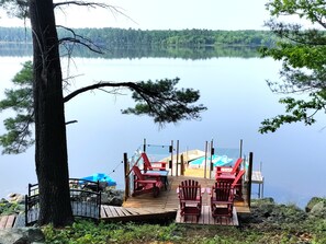 A dock with chairs and a small boat, overlooking a calm lake with trees in the background.