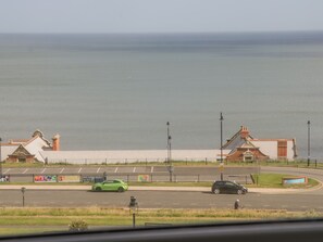 A coastal road with a green car and a black car, a pedestrian, and a building with a red roof. - Foxton Penthouse (Whitby)