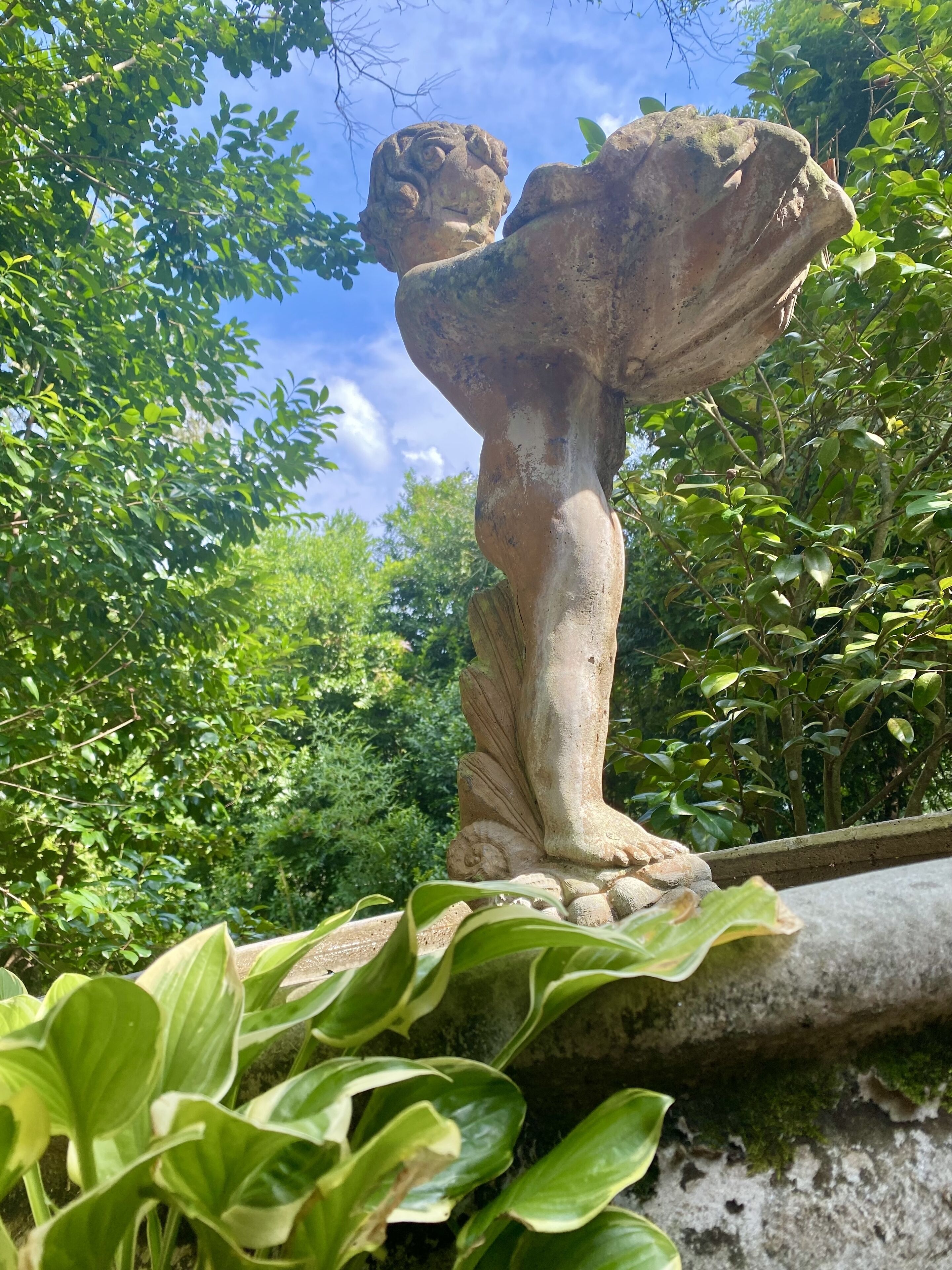 A stone statue of a figure with a leaf-like structure on its head, surrounded by lush greenery.