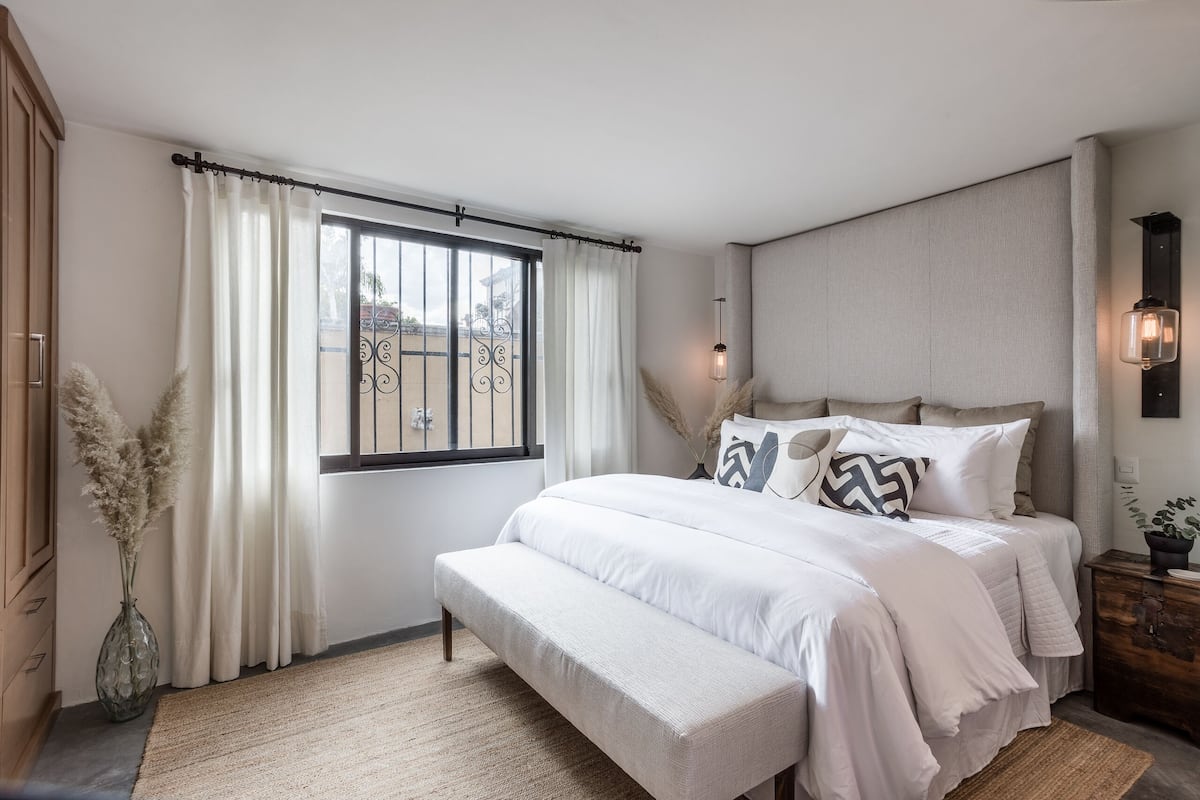 Een slaapkamer met een groot bed, een bankje, een raam met gordijnen en een nachtkastje met een lamp.