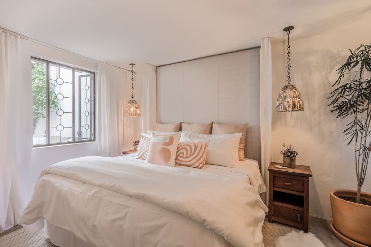 Een slaapkamer met een groot bed, nachtkastjes, een kamerplant en een raam met gordijnen.