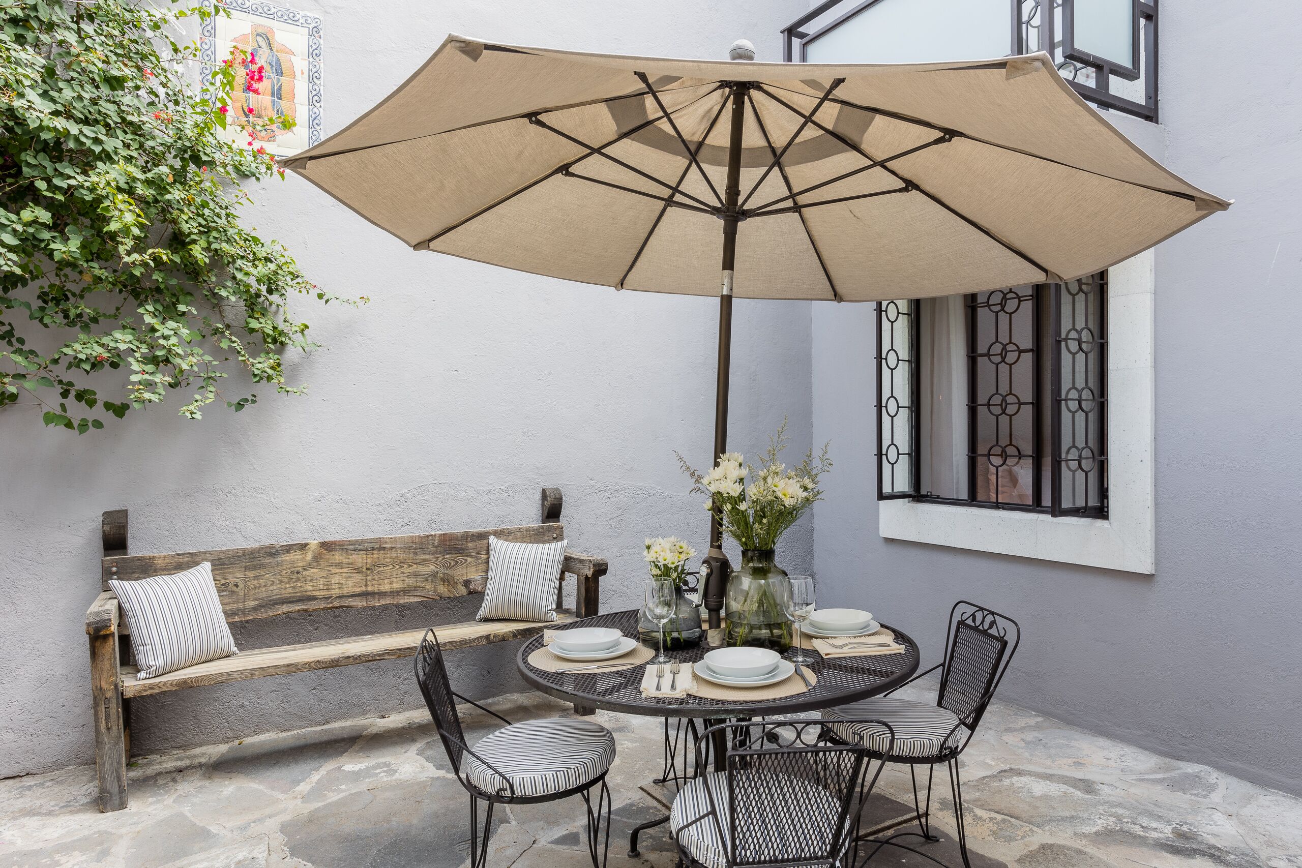Une terrasse avec une table ronde, des chaises, un banc, un parasol et un vase rempli de fleurs.