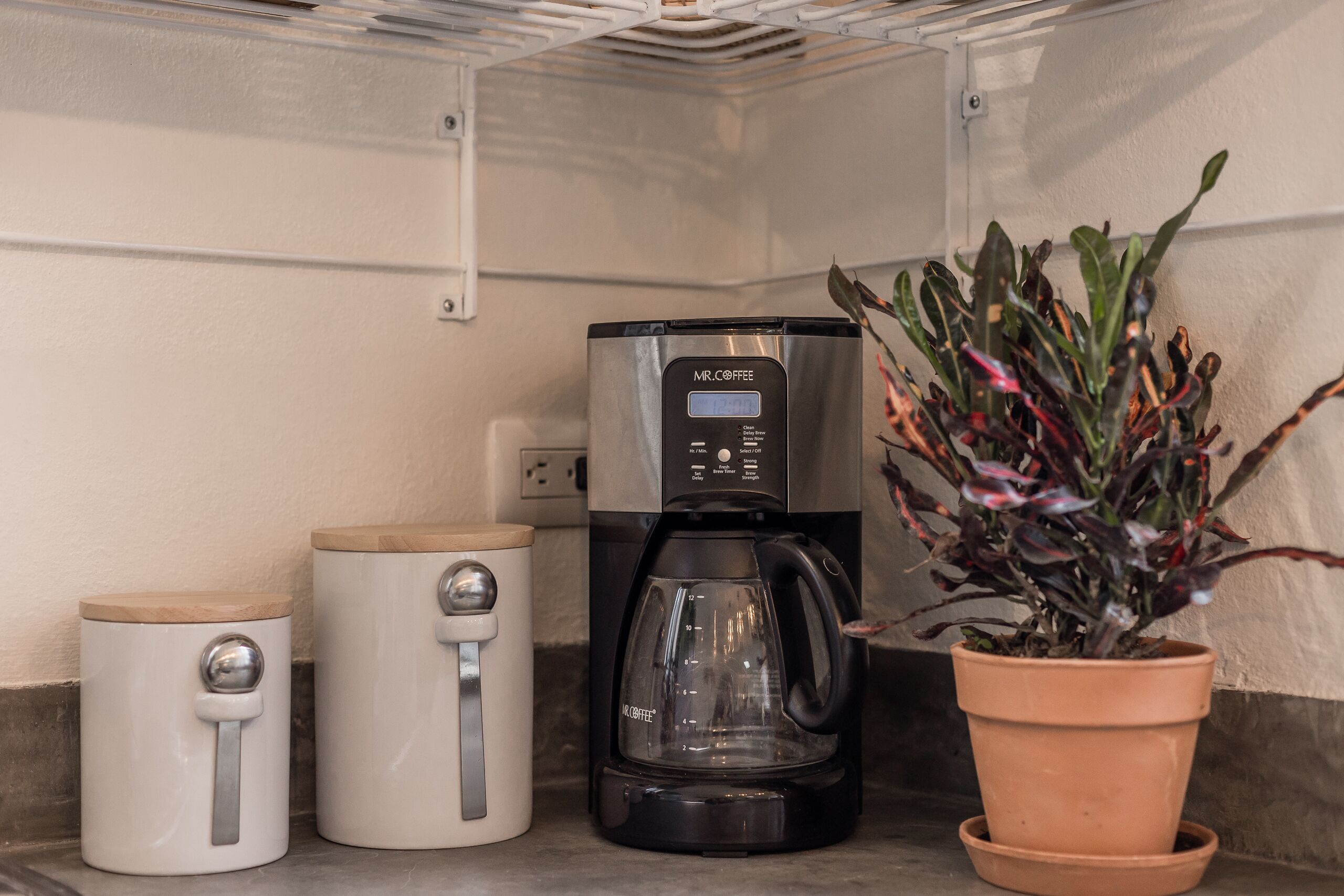 A compact kitchen area with a Mr. Coffee espresso machine, a white coffee maker, and a potted plant.