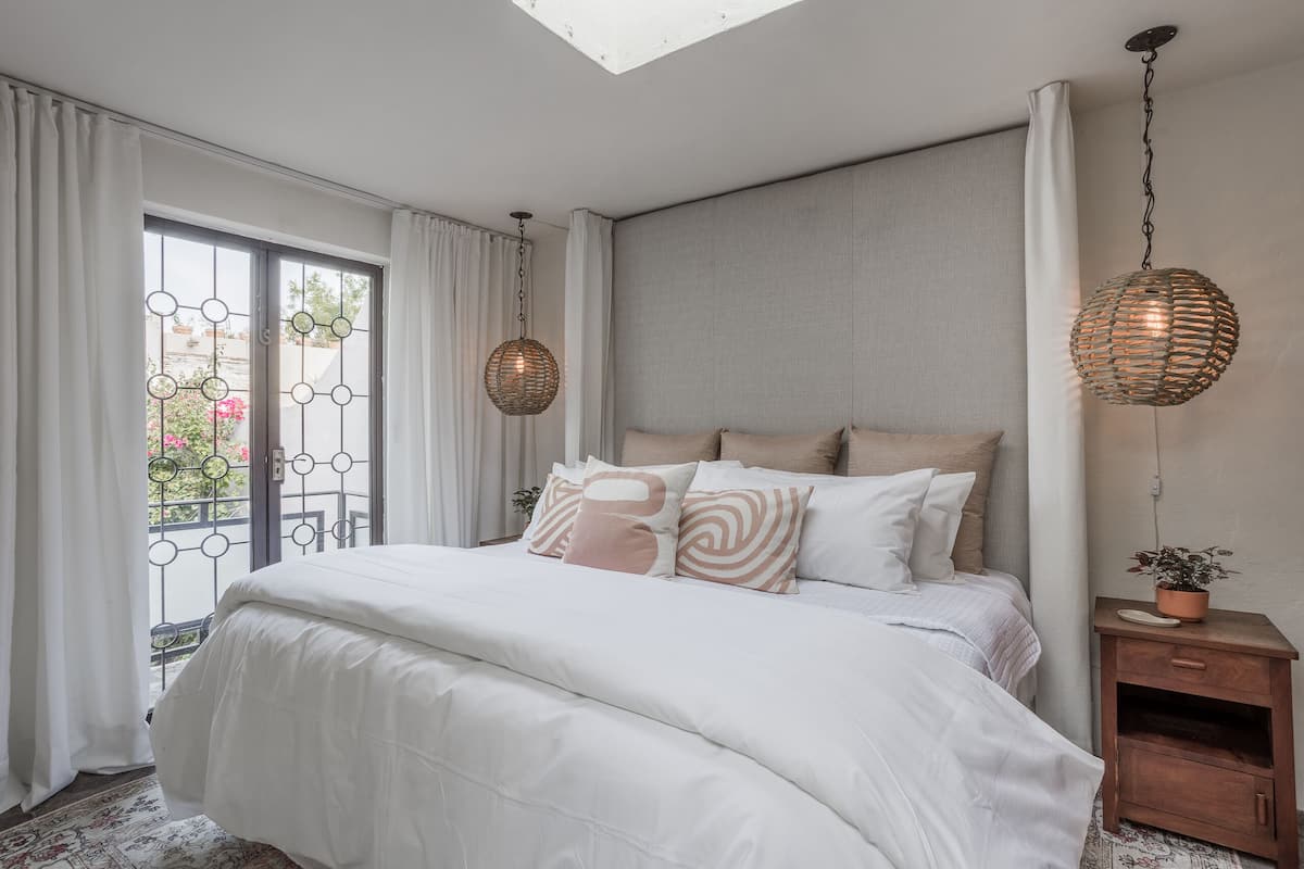Een slaapkamer met een groot bed, een nachtkastje met een plant en uitzicht door een raam met gordijnen.
