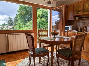 A dining area with a table and chairs, a bowl of fruit, and a view of greenery outside. - Vintage (Annecy-le-Vieux)