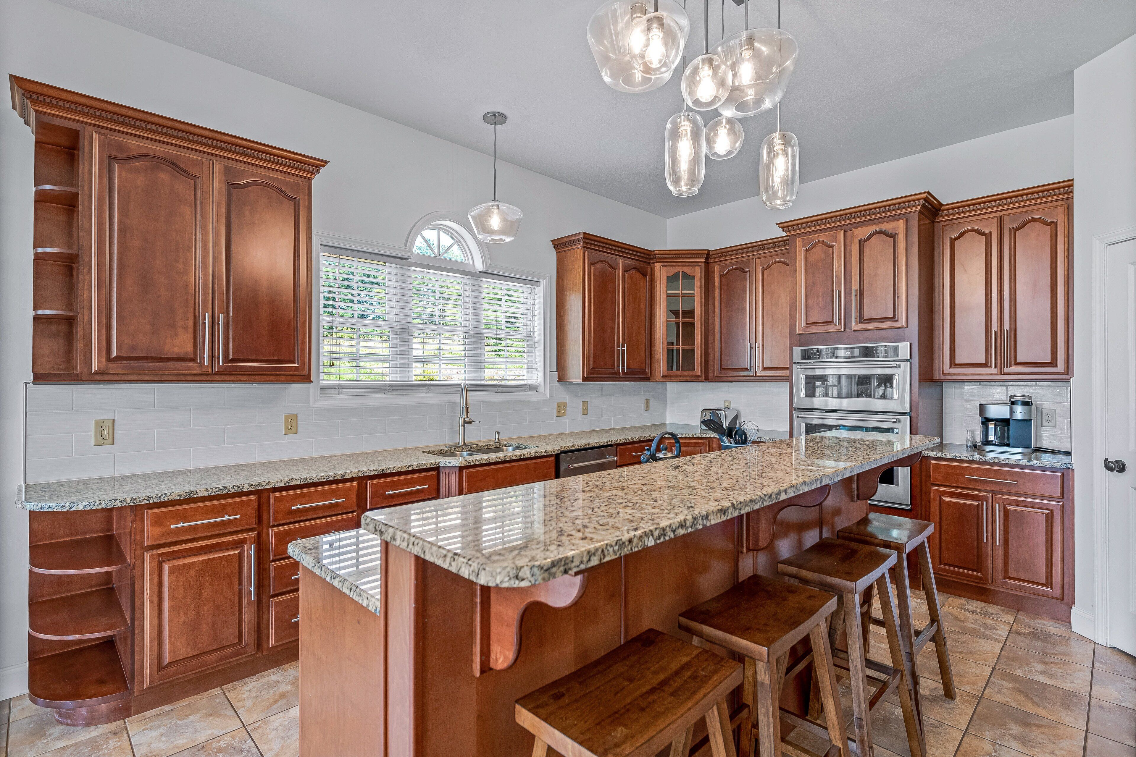 A kitchen with wooden cabinets, granite countertops, a large island with stools, stainless steel appliances, and pendant lighting.