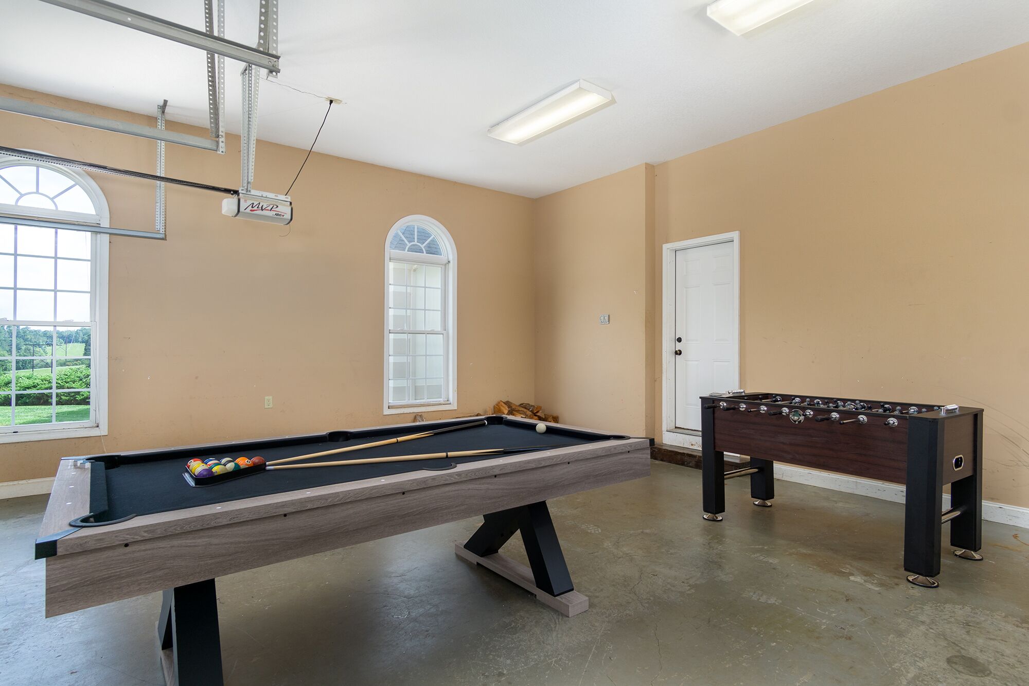 A pool table with a billiard cue and balls, a foosball table, and a window with a view of greenery.