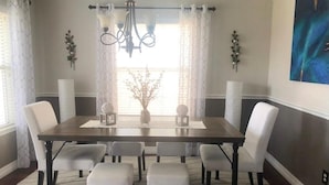 Comedor con una mesa rectangular, seis sillas blancas tapizadas, una araña y un amplio ventanal con cortinas traslúcidas.