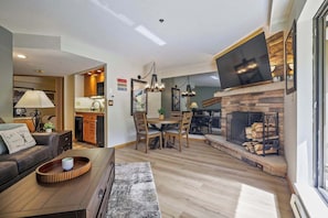 A modern living room with a fireplace, a dining area, and a kitchen visible through an open door.