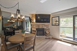 A dining area with a round table and chairs, a fireplace, a TV mounted on the wall, and a large window with a view of the outdoors.