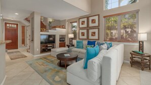 A spacious living room with a white sofa, a coffee table, and a TV. There are framed pictures on the wall and a window with blinds.