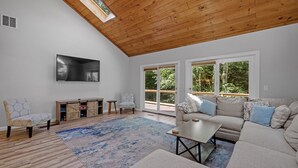 A spacious living room with a high ceiling, a large flat-screen TV mounted on the wall, a beige sectional sofa, a coffee table, and a patterned rug. - Spacious home minutes from Gunstock and Lake Winnipesaukee (Gilford)