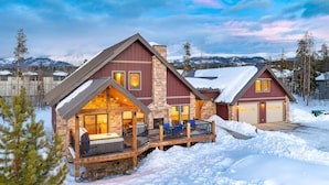Une grande cabane en rondins à deux étages, dotée d’une terrasse et d’une cheminée, entourée de neige.