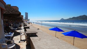 Una playa con tumbonas y sombrillas, una red de voleibol y vistas al océano y las montañas.
