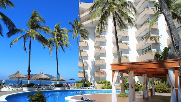 Un área de piscina en un resort con palmeras, sillas de descanso y un edificio con balcones.