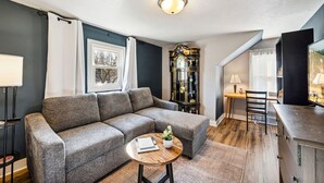 Un salón con un sofá seccional gris, una mesa de centro redonda de madera, un mueble negro y una ventana con cortinas blancas.