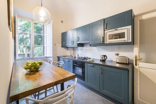 A kitchen with blue cabinets, a wooden dining table, and a window with a view of greenery.