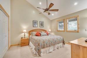 Quarto com cama, mesas de cabeceira, ventilador de teto e duas janelas com persianas.
