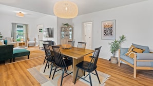 A modern living room with a wooden dining table, black chairs, a large woven pendant light, and a grey sofa with a blue cushion.