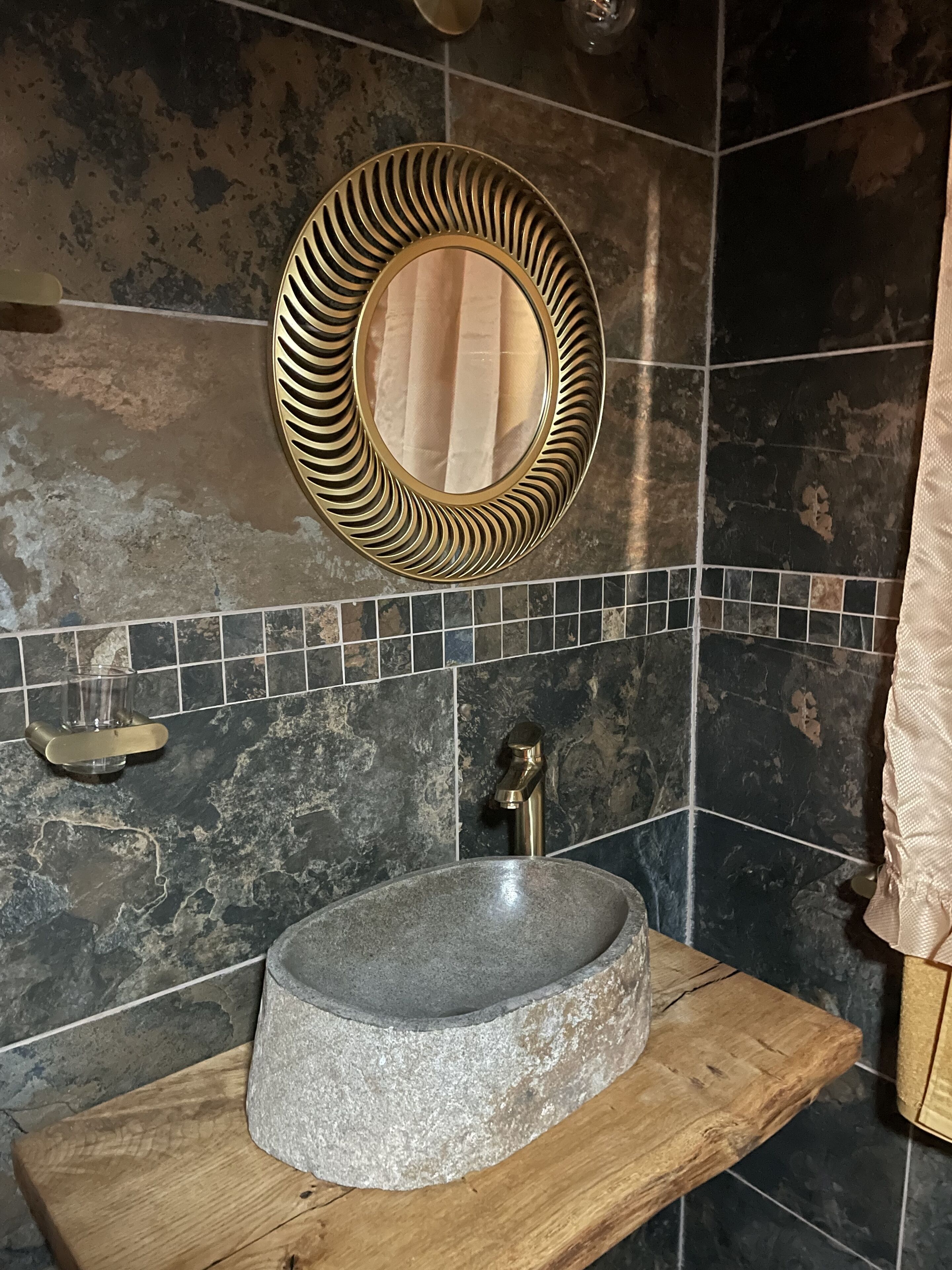 A bathroom with a stone sink, a gold-framed mirror, and a tiled backsplash.