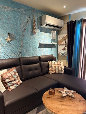 A modern living room with a black leather sofa, patterned cushions, a wooden coffee table, and a decorative wall with a blue textured pattern and wall-mounted air conditioning unit.