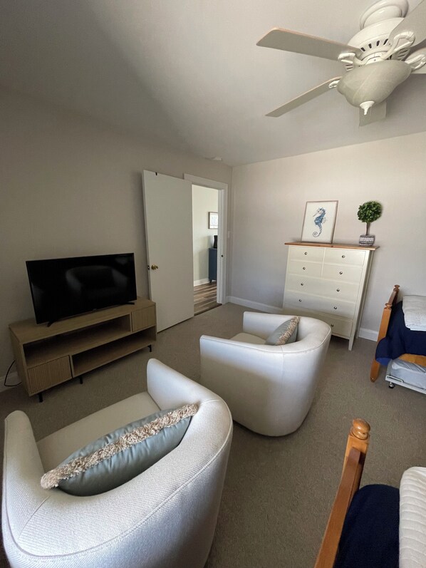 A living room with a flat-screen TV, two white armchairs, a wooden TV stand, a white dresser, and a ceiling fan.