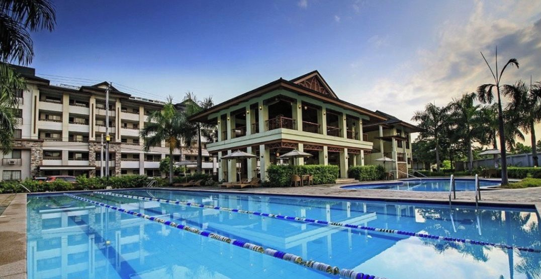 A swimming pool with clear blue water and lane dividers, surrounded by a building with multiple balconies and a tropical setting.