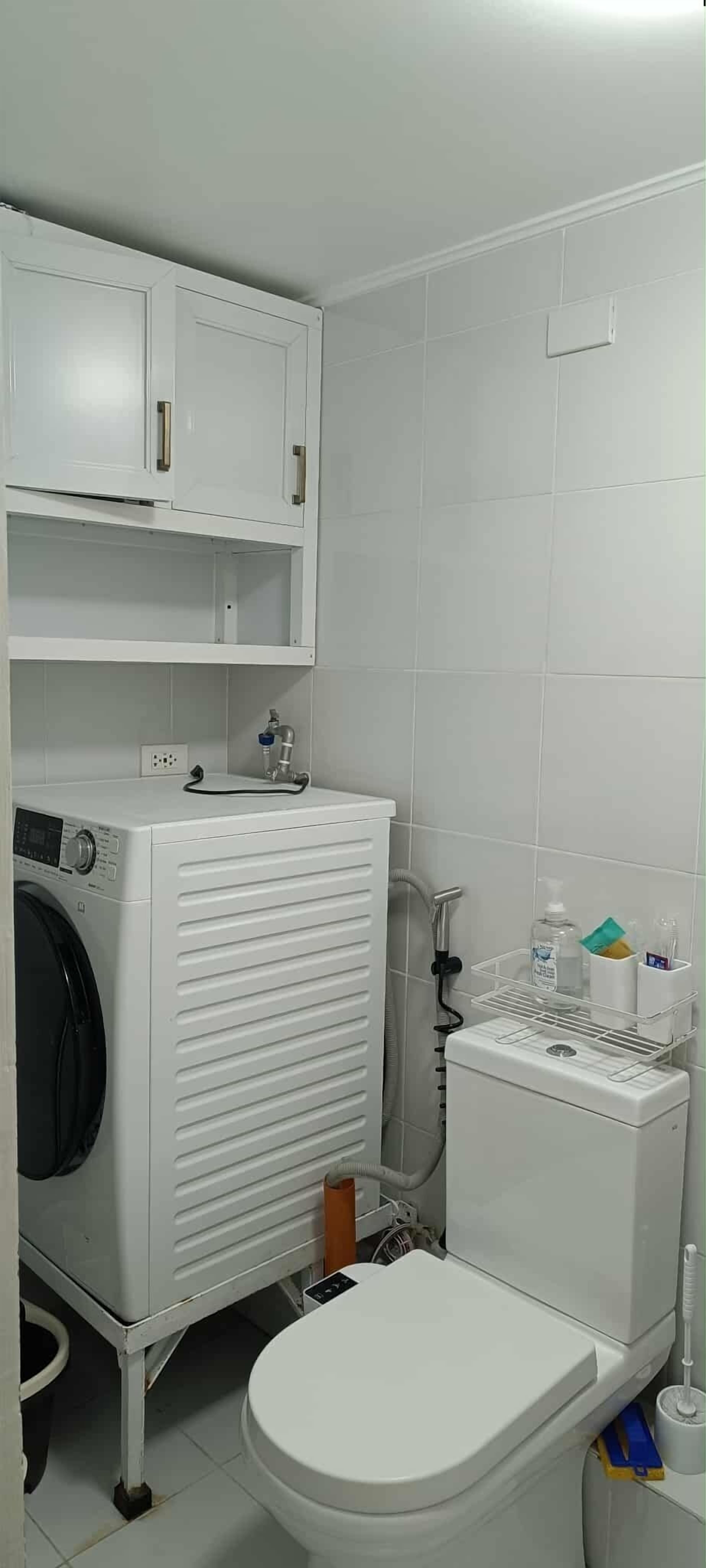 A compact bathroom with a washing machine, toilet, and a shelf with toiletries.