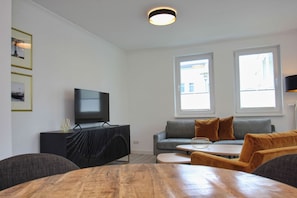 A modern living room with a flat-screen TV on a black entertainment unit, a grey sofa with orange cushions, a wooden coffee table, and two windows providing natural light.