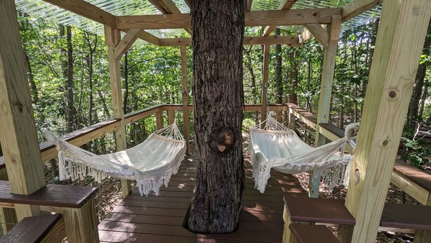 Un kiosque en bois avec un tronc d’arbre au centre, entouré d’une terrasse en bois dotée de bancs et de deux hamacs.