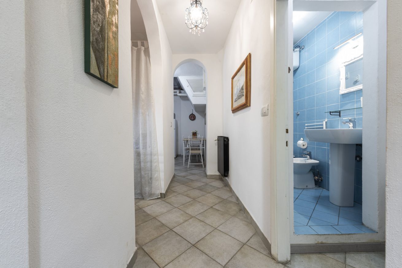 Ein Flur mit Fliesenboden, einem gerahmten Bild an der Wand und Blick in ein Badezimmer mit blauen Fliesen und einem Waschbecken.