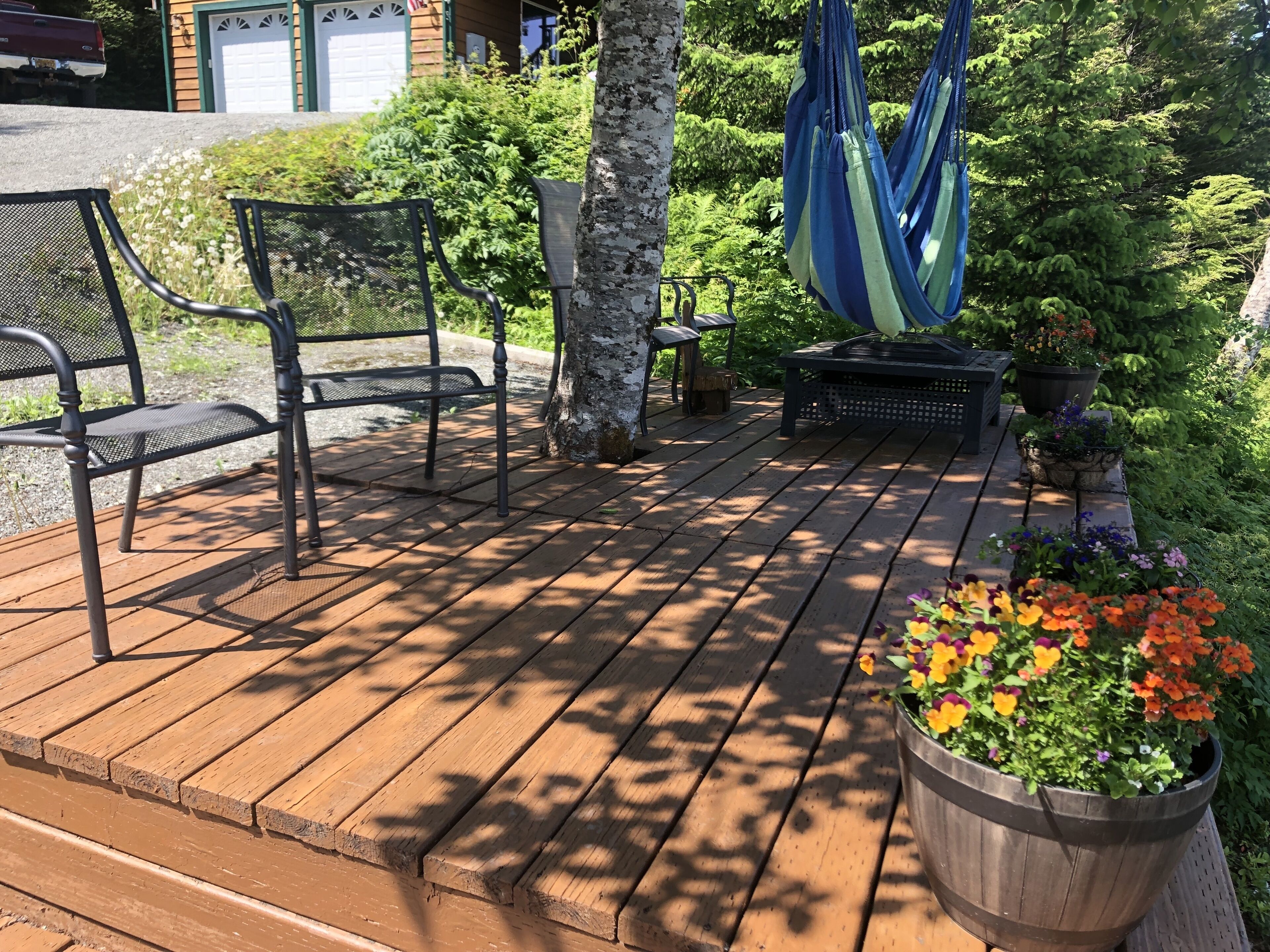 A wooden deck with outdoor furniture, including chairs and a hammock, surrounded by greenery and potted plants.