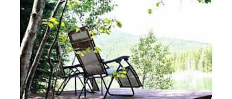 Una terraza de madera con una silla metálica y una mesa, con vista a un lago y colinas boscosas.