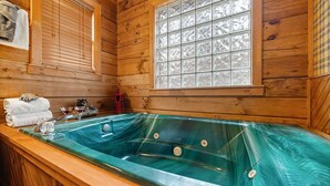 Interno di una baita in legno con vasca idromassaggio in vetro, pareti di legno, una finestra con vetro smerigliato e un porta asciugamani.