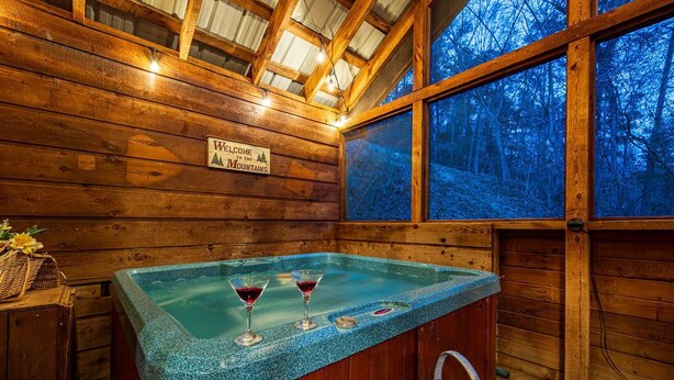 Interno di una baita in legno con una vasca idromassaggio, due bicchieri di vino rosso e un cartello di benvenuto.