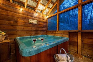 A wooden cabin interior with a hot tub, two glasses of red wine, and a welcome sign.
