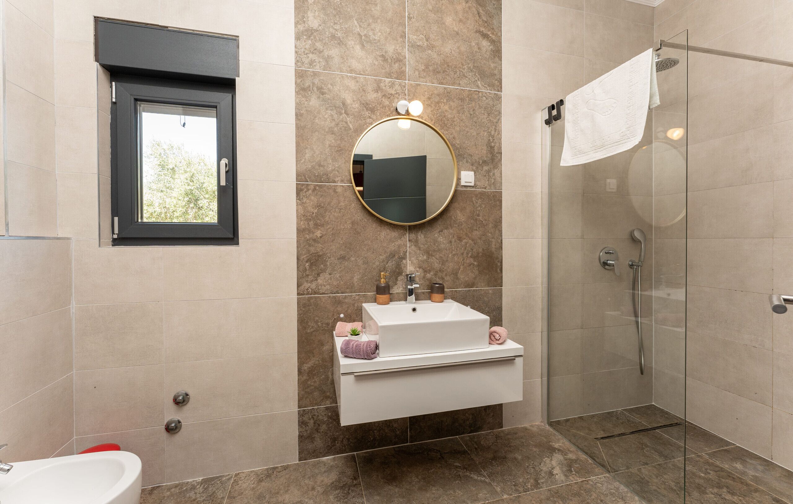 Un baño moderno con cabina de ducha de vidrio, un lavabo blanco con espejo encima y una ventana con marco negro.