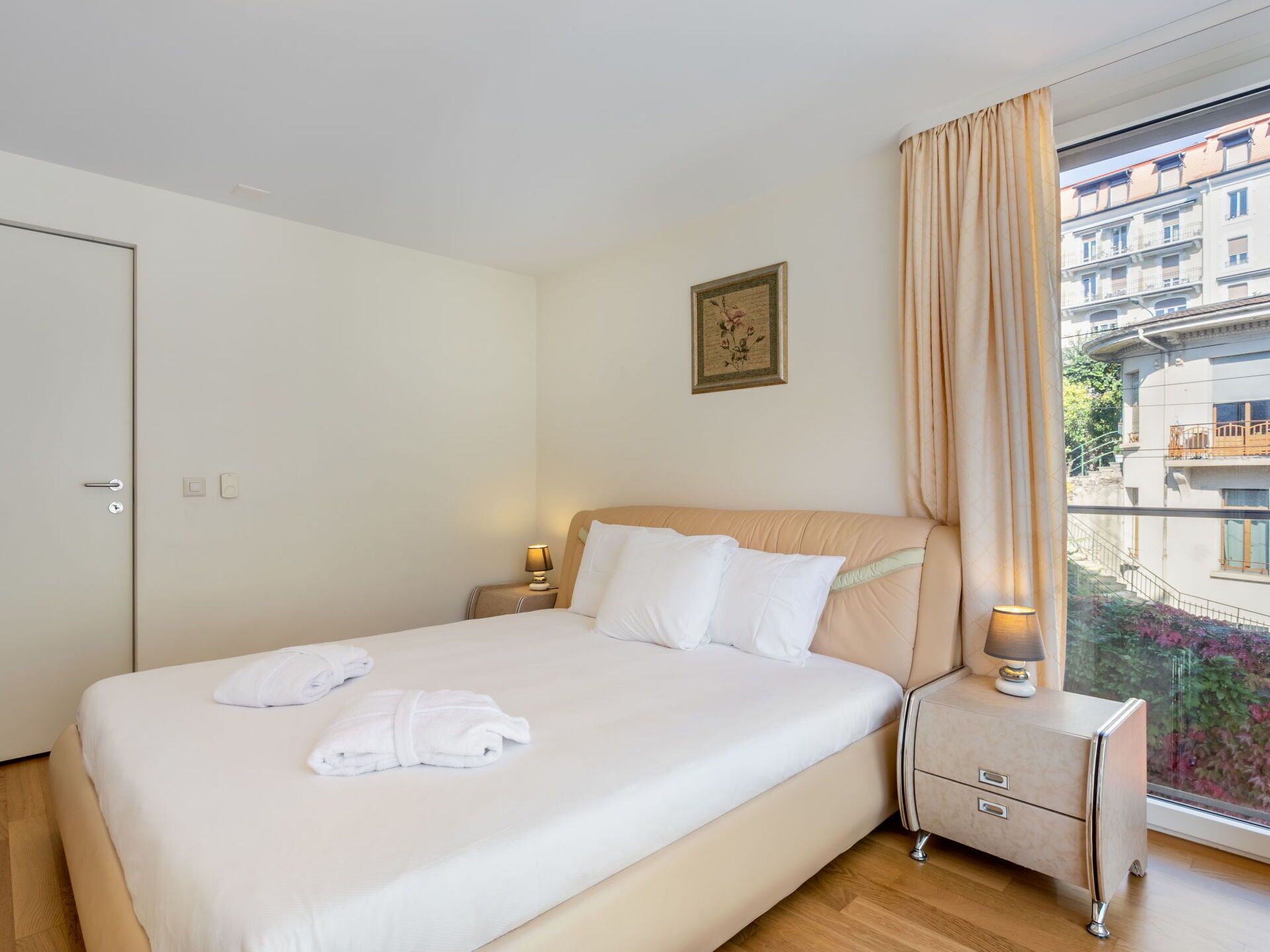 Habitación de hotel con cama, mesitas de noche, ventana con cortinas y vista a edificios.