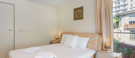 Habitación de hotel con cama, mesitas de noche, ventana con cortinas y vista a edificios.