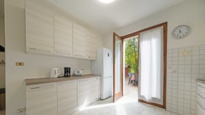 A modern kitchen with light wood cabinets, a white refrigerator, a coffee maker, and a toaster. There is a clock on the wall and a door leading to an outdoor area with chairs. - Casa Ruby - Pool and tennis court Lazise/Pacengo (Lazise)