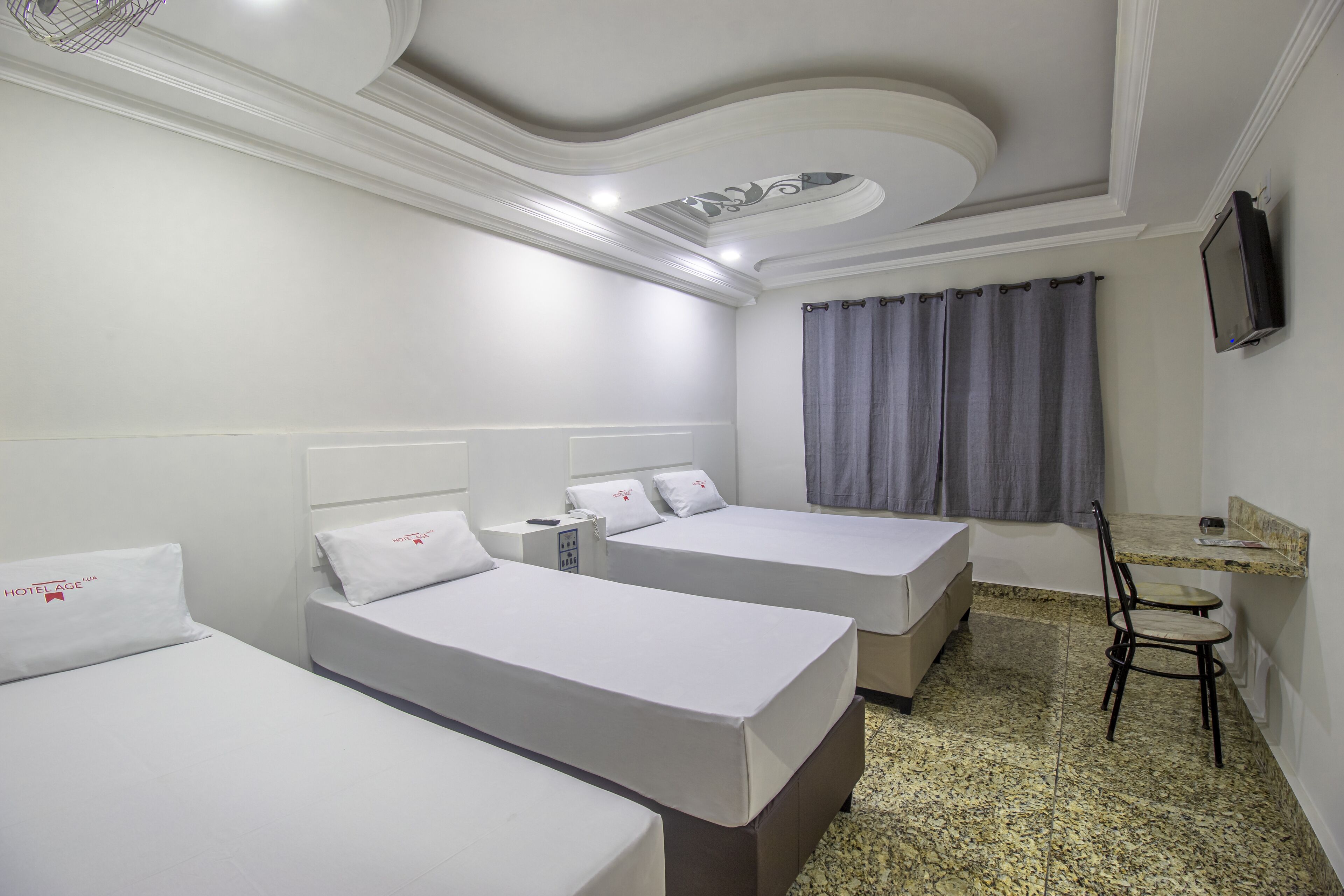 A hotel room with three single beds, a small desk with a chair, a television on the wall, and a window with curtains.