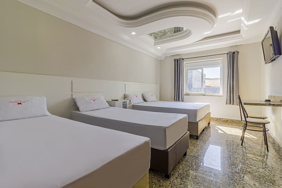 A hotel room with three beds, a small table, a chair, a TV, and a window with curtains.