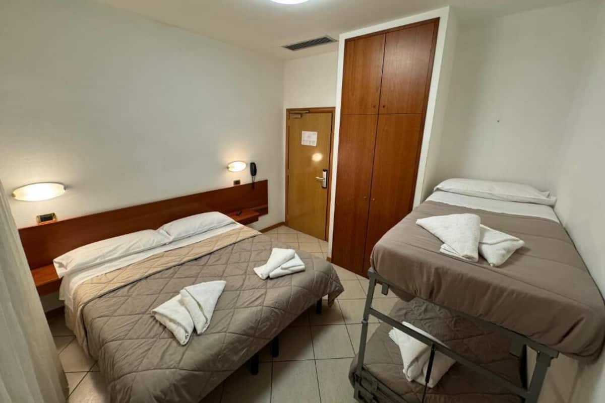 A hotel room with two beds, a wooden wardrobe, and a bedside table with a lamp.