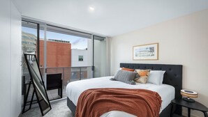 Une chambre moderne avec un grand lit, une petite table de chevet, un tableau encadré au mur et une vue sur l’extérieur grâce à une porte coulissante.