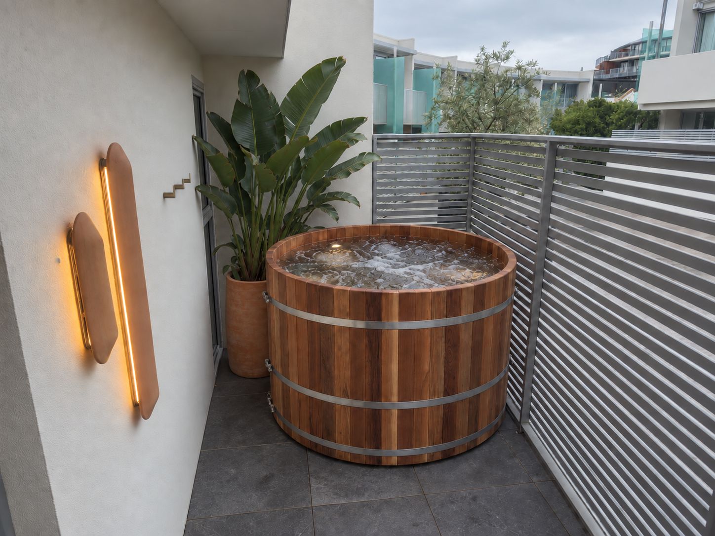 A wooden hot tub on a balcony with a potted plant and a wall-mounted light fixture.