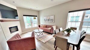A modern living room with a sofa, coffee table, and dining table. There is a fireplace, large windows, and a sign on the wall. - Luxury 3BR/4BA Townhome • Rooftop Deck, Porch • 5 Min to Downtown (Omaha)