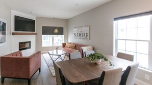 A modern living room with a wooden dining table, cushioned seating, a fireplace, and large windows. - Luxury 3BR/4BA Townhome • Rooftop Deck, Porch • 5 Min to Downtown (Omaha)