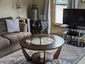 A living room with a round wooden coffee table, a sofa, a vintage trunk, a television on a stand, and a window with curtains.