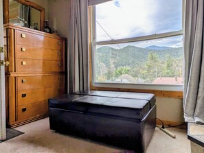 A room with a wooden dresser, a black ottoman, and a window with a view of a mountainous landscape.