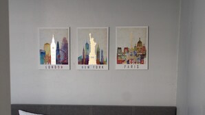Un dormitorio con una cama, almohadas y una pared con tres carteles de paisajes urbanos que muestran Londres, Nueva York y París.